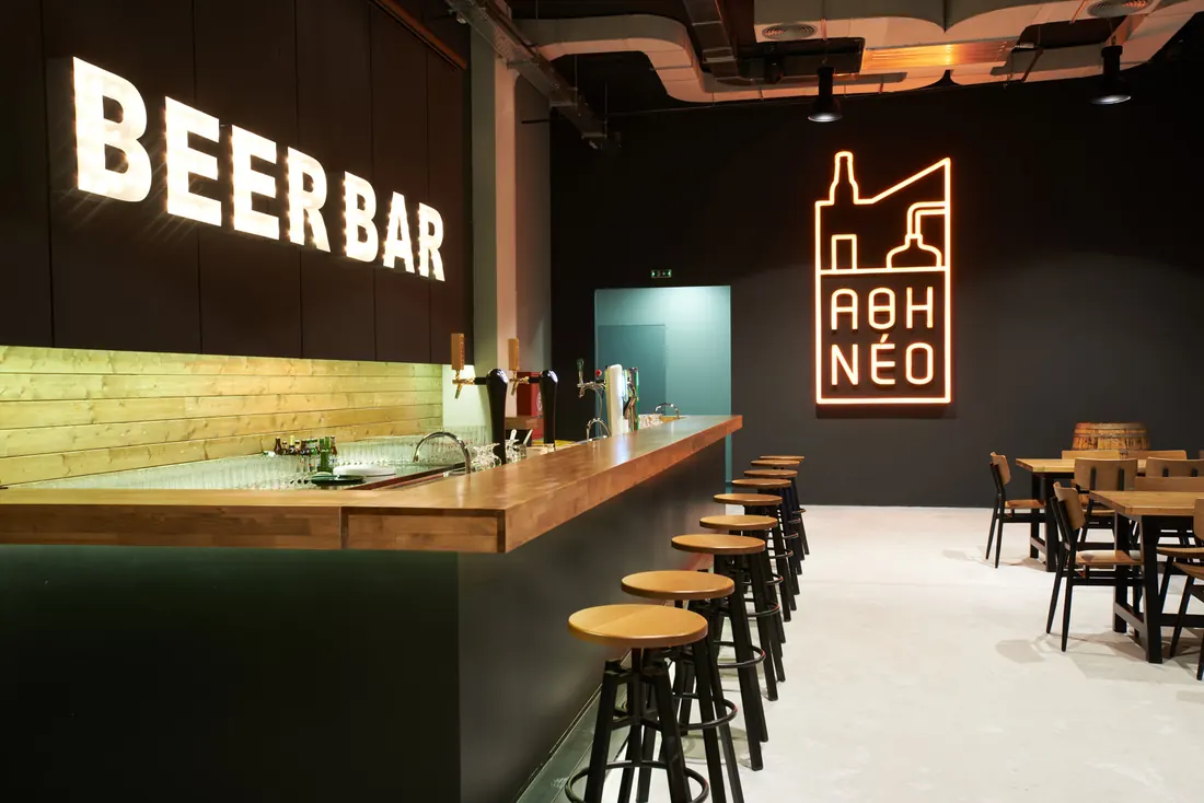 Το Beer Bar του ΑΘΗΝΕΟ με ξύλινο πάγκο, σκαμπό και φωτεινή επιγραφή